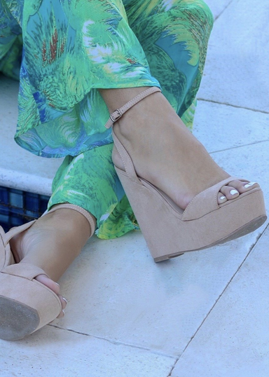 Nude suede ALYSSA wedge with crisscross strap that crosses in the back as the buckle buckles in the side that is silver. Thick to thin back to thick strap in the front. Mode is wearing green tropical pants on a stone  floor.