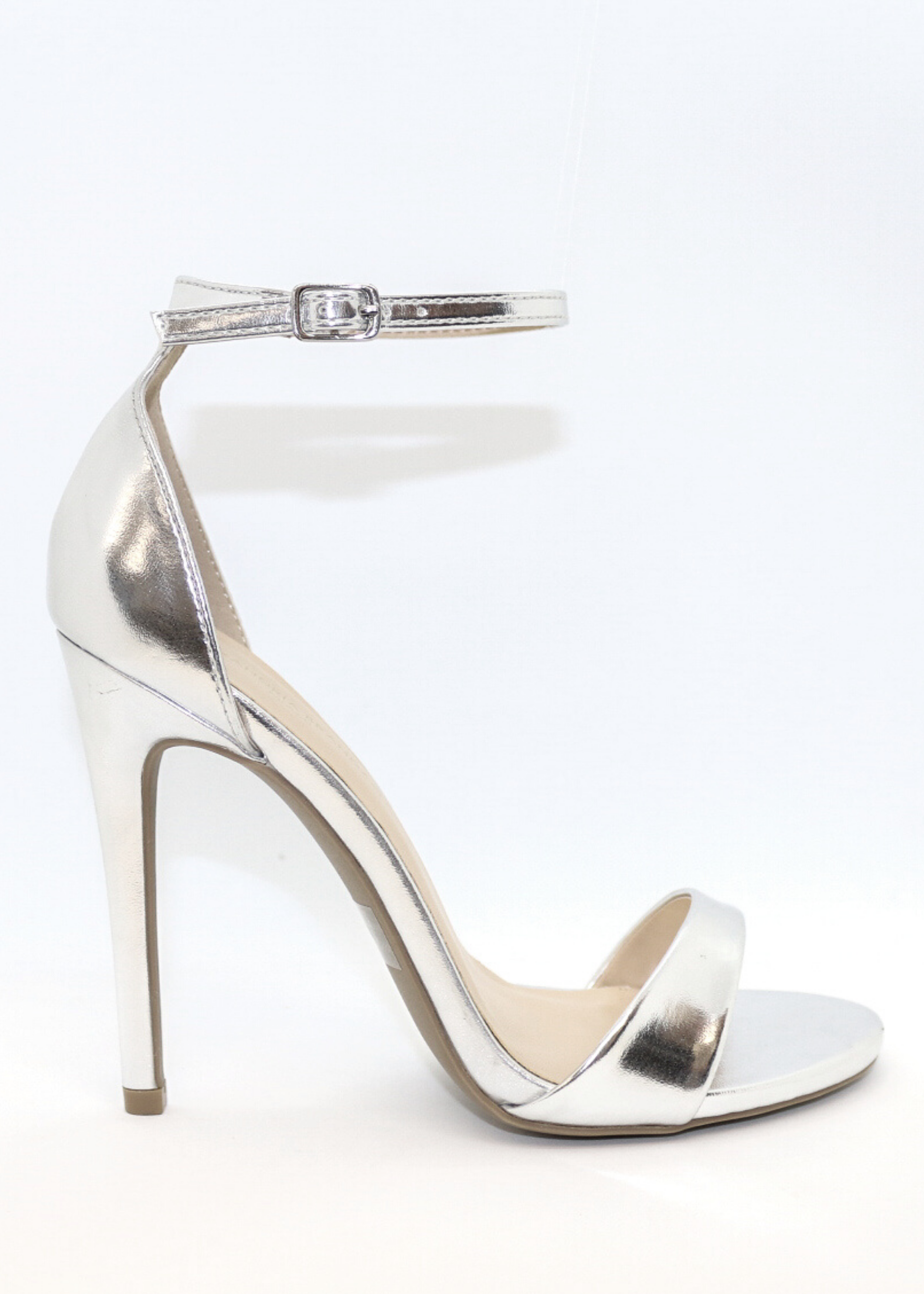 Ali Heels in Silver as the feet is propped on a marble table is the back ground. It is pictured on a side view as you see buckle around the ankle. Ali Heels in Silver is a heel that is covered in the back of the heel and has a thick to thin to thick strap in the front showing your toes.