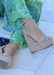 Nude suede ALYSSA wedge with crisscross strap that crosses in the back as the buckle buckles in the side that is silver. Thick to thin back to thick strap in the front. Mode is wearing green tropical pants on a stone  floor.