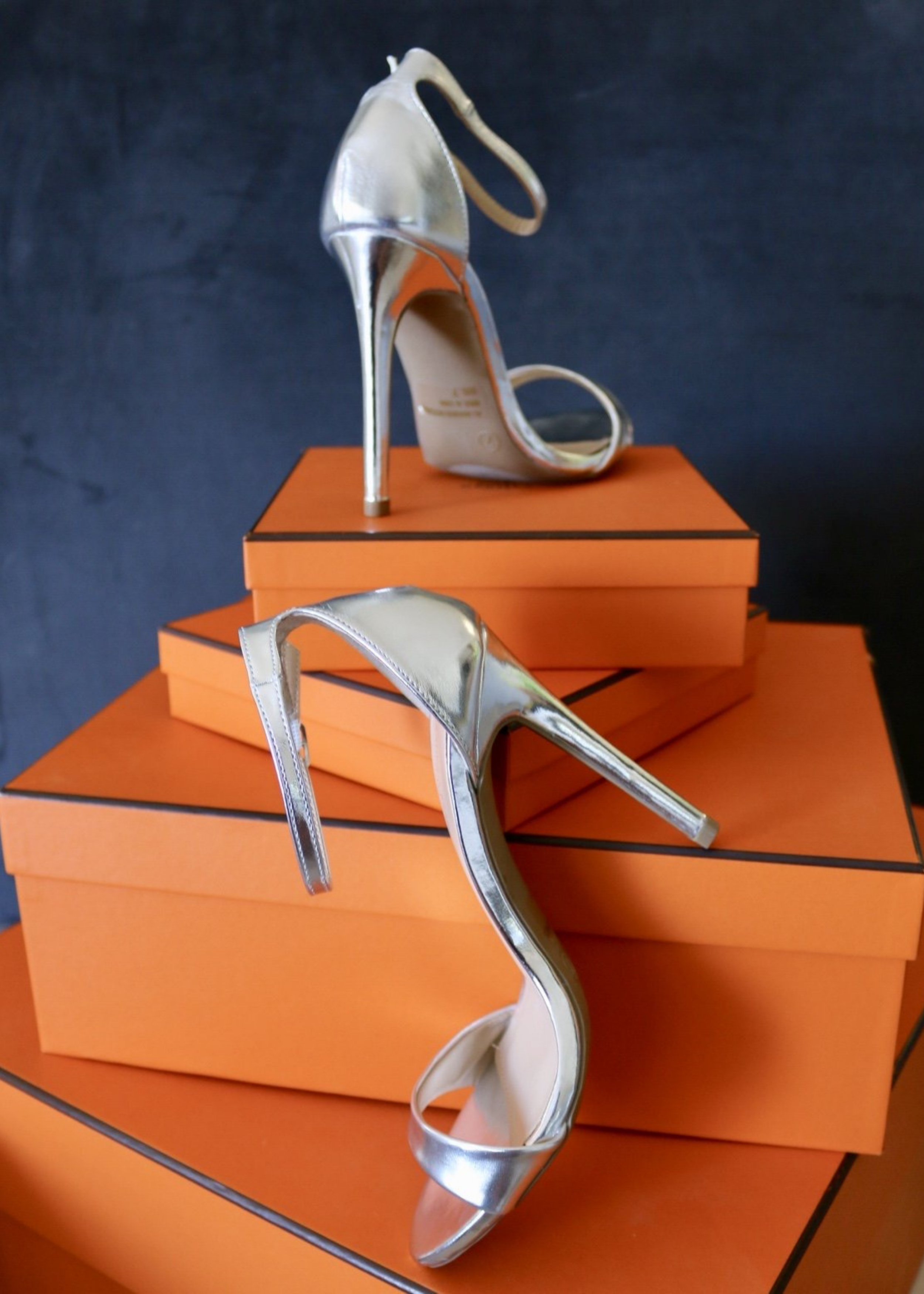 Silver Metallic "Ali" sandal heels propped up on orange boxes.