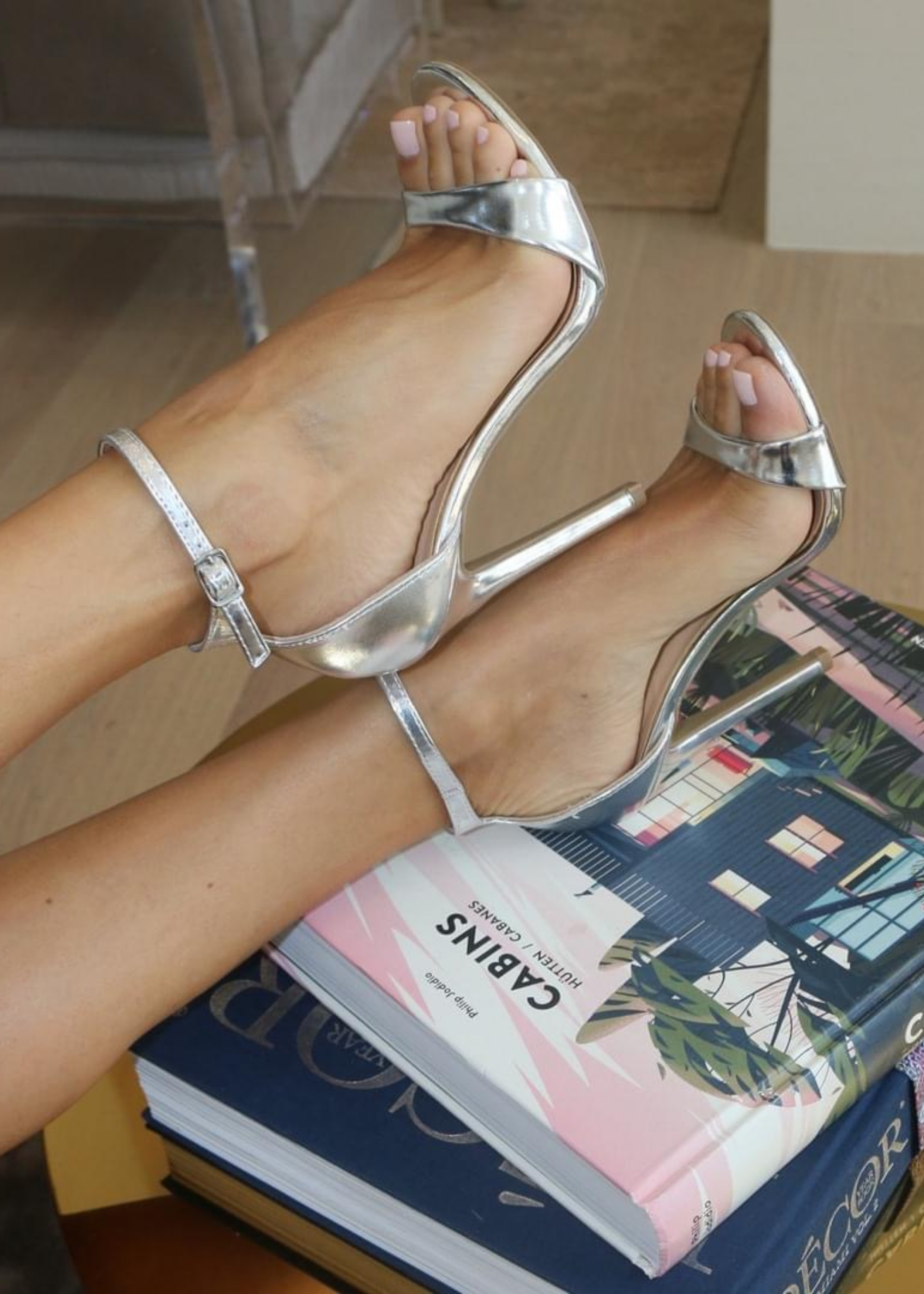 Ali Heels in Silver as the feet is propped on colorful books on a  table and candles in the back ground. It is pictured on a side view as you see buckle around the ankle. Ali Heels in Silver is a heel that is covered in the back of the heel and has a thick to thin to thick strap in the front showing your toes.